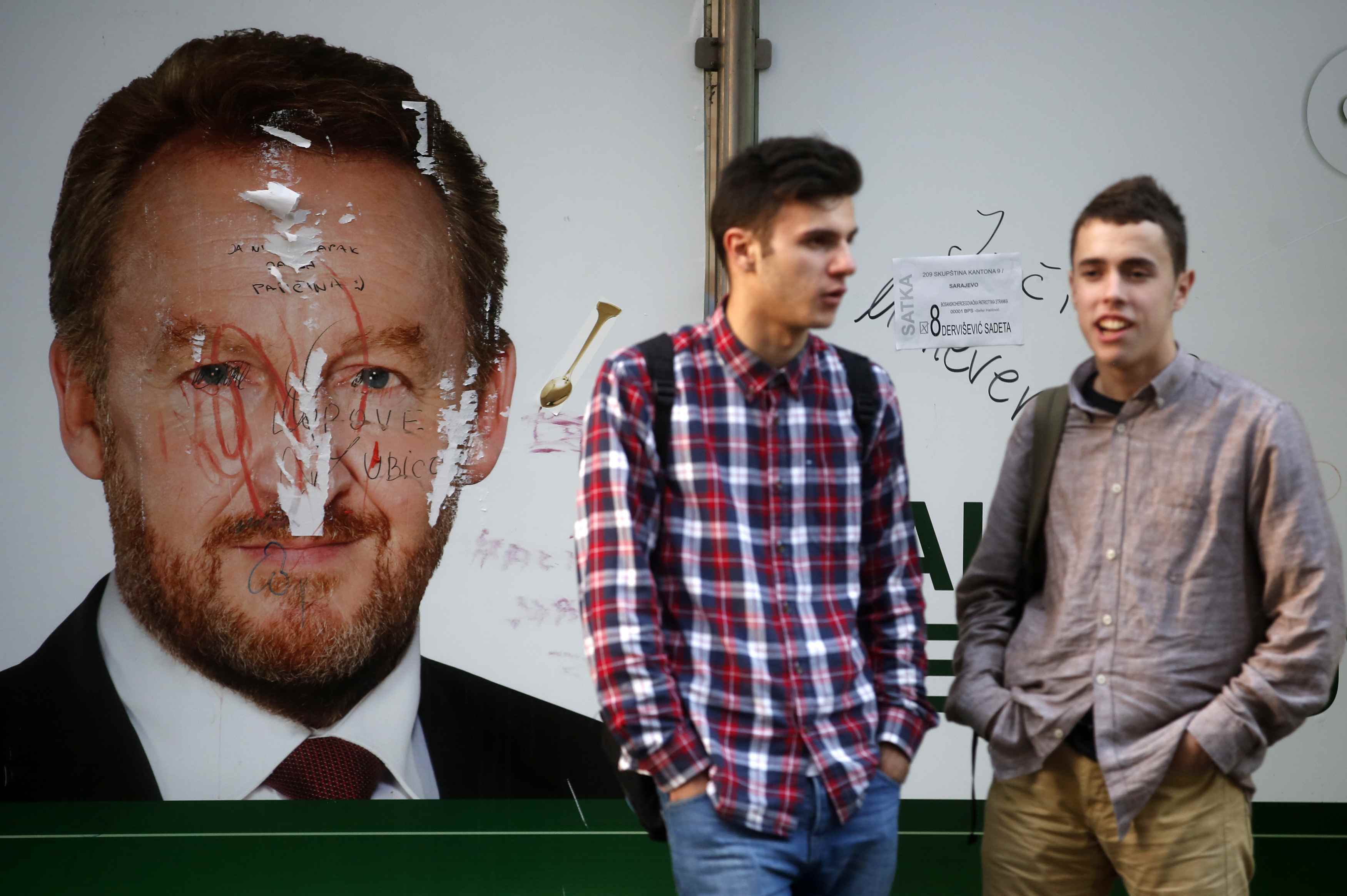 Men stand at a bus station in front of a defaced election poster in Sarajevo, October 10, 2014. Bosnians will vote on Sunday for a parliamentary and presidential election that could break the reform deadlock in the ethnically divided country or deepen div