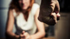 Young woman is sitting hunched at a table at home, the focus is on a man's fist in the foregound of the image